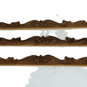 Puede incluir: Tres molduras decorativas de madera con acabado marrón oscuro. Cada pieza presenta un diseño floral y de volutas repetido a lo largo del borde superior. La moldura es probablemente para muebles o detalles arquitectónicos.