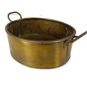 Op de afbeelding: Een vintage koperen plantenbak met twee handvatten. De plantenbak heeft een versleten, verweerde uitstraling en is ovaal.