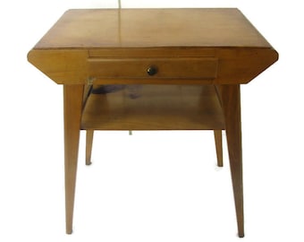 May include: A vintage wooden side table with a drawer and a lower shelf. The table has a light brown finish, a small drawer with a dark knob, and tapered legs. The table measures approximately 24 inches wide, 20 inches deep, and 24 inches high.