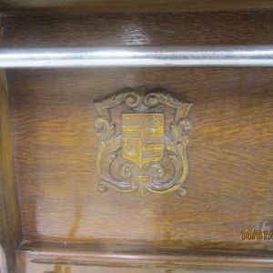 May include: A wooden cabinet with a carved crest and a metal bar. The cabinet is made of dark wood and has a mirror on the right side. The crest is in the center and has a shield design.