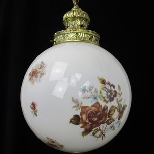 May include: A vintage white glass pendant light with a floral design. The globe-shaped lamp features a brass-colored crown and base. The floral pattern includes roses in shades of brown, pink, and blue.