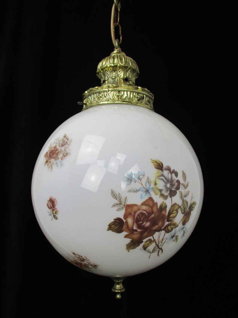 May include: A vintage white glass pendant light with a floral design. The globe-shaped lamp features a brass-colored crown and base. The floral pattern includes roses in shades of brown, pink, and blue.