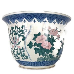 Op de afbeelding: Een witte keramische plantenpot met blauwe en groene bloemenmotieven. De pot heeft een brede rand en een afgeronde basis.