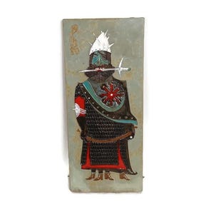 Op de afbeelding: Een keramische wandplaat met een gestileerde krijgerfiguur in zwart harnas met een rood-witte band. De krijger draagt een witte helm met een puntige kam en houdt een zwaard in zijn mond. De achtergrond is lichtblauwgroen.