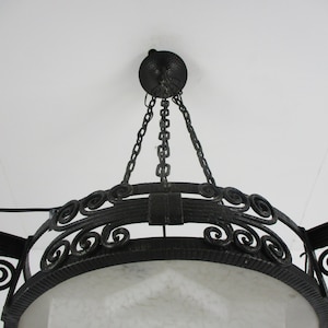 May include: An ornate, wrought-iron chandelier with a circular design. The fixture is suspended from the ceiling by chains and features decorative scrollwork and a textured, dark finish. The light fixture has a white, frosted glass shade.