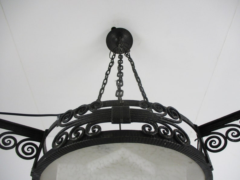 May include: An ornate, wrought-iron chandelier with a circular design. The fixture is suspended from the ceiling by chains and features decorative scrollwork and a textured, dark finish. The light fixture has a white, frosted glass shade.