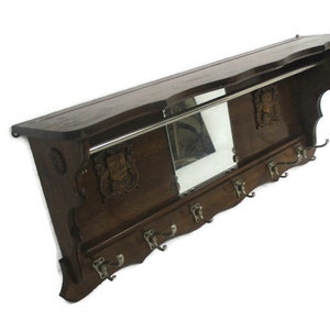 May include: A dark brown wooden wall-mounted coat rack with a shelf, mirror, and coat hooks. The rack features decorative carvings and a metal bar for hanging items. The hooks are silver-toned.