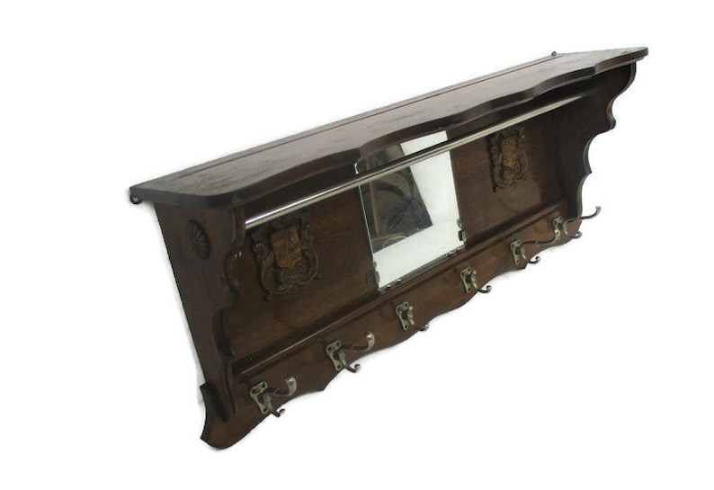 May include: A dark brown wooden wall-mounted coat rack with a shelf, mirror, and coat hooks. The rack features decorative carvings and a metal bar for hanging items. The hooks are silver-toned.