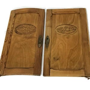 May include: Two antique wooden cabinet doors. Each door is a light brown color with a carved oval floral design. The doors have brass hardware and are set against a white background. The doors are likely from a vintage cabinet.