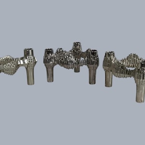 May include: Three silver-colored metal candle holders, each featuring four cylindrical legs and a textured, jagged, architectural-style frame connecting the top sections. The metallic surfaces have a reflective finish with intricate, vertical ridged detailing.