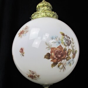May include: A vintage white globe pendant light with floral rose and blue flower designs. The light fixture has a brass-colored top and bottom, and is suspended by a chain.