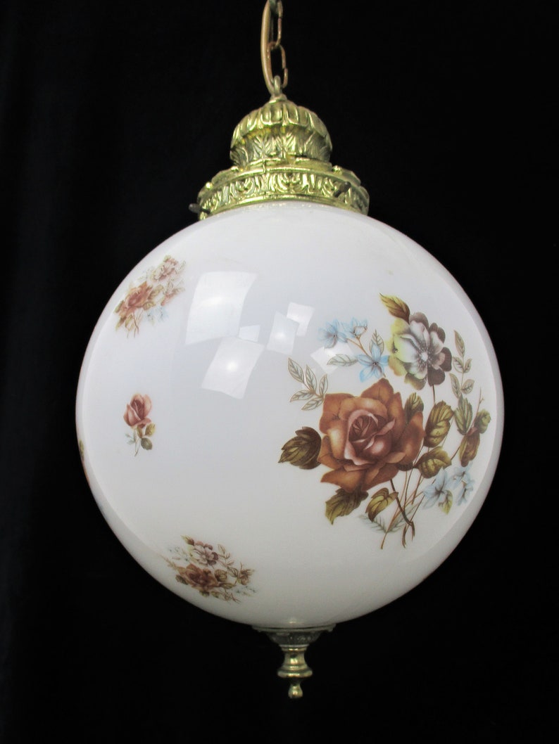 May include: A vintage white globe pendant light with floral rose and blue flower designs. The light fixture has a brass-colored top and bottom, and is suspended by a chain.