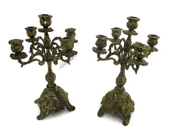 May include: Two antique brass candelabras with five candle holders each. The candelabras have ornate, swirling designs and sit on a decorative base. The brass has a warm, aged patina, suggesting vintage craftsmanship.