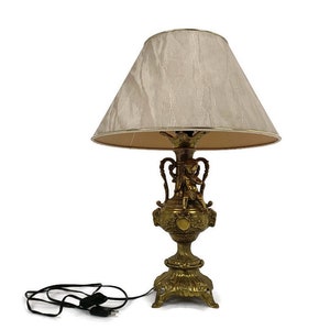 May include: A gold table lamp with a white lampshade. The lamp base features a sculpted cherub design.