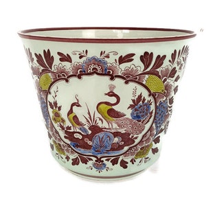 Op de afbeelding: Een witte keramische plantenpot met een bruin, blauw en geel bloemenontwerp met pauwen. De pot heeft een brede, ronde basis en een licht uitlopende rand.