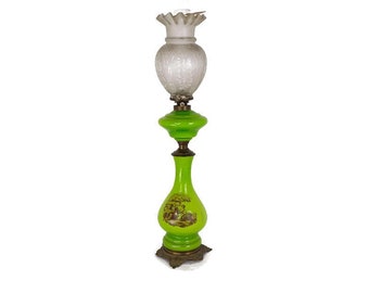 May include: A tall green glass oil lamp with a white glass shade. The lamp has a decorative design on the base and a floral design on the shade.