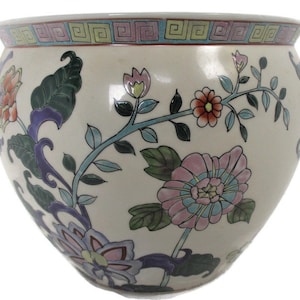 May include: A white ceramic planter with a floral design. The planter features pink, blue, green, and orange flowers with green leaves. The planter has a wide, round base and a narrow opening.