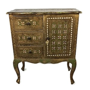 May include: A small, antique-style wooden cabinet with three drawers and a single door. The cabinet is painted in a gold and green color scheme with intricate floral and geometric patterns. The cabinet has four legs and is made of wood.
