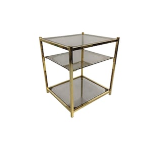 Puede incluir: Una mesa auxiliar de tres niveles con estructura de metal dorado y estantes de vidrio transparente. La mesa tiene forma cuadrada y está diseñada con una estética minimalista. Los estantes de vidrio están espaciados uniformemente.