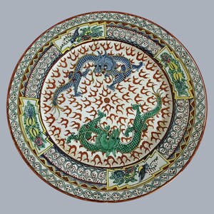 Könnte beinhalten: Ein runder Keramikteller mit einem zentralen Motiv aus einem blauen und einem grünen Drachen, umgeben von roten Flammenmotiven. Der Rand ist mit geometrischen Mustern, floralen Elementen und stilisierten Formen in Gelb, Grün und Blau verziert.