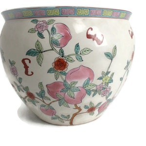 Puede incluir: Un macetero de cerámica blanca con un diseño floral. El macetero presenta flores rosas y rojas, hojas verdes y fruta de color melocotón. El borde del macetero está decorado con un patrón geométrico azul y verde.