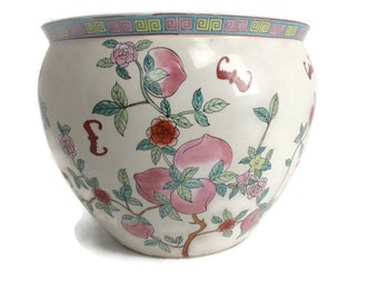 Könnte beinhalten: Ein weißer Keramiktopf mit Blumenmuster. Der Topf zeigt rosa und rote Blumen, grüne Blätter und pfirsichfarbene Früchte. Der Rand des Topfes ist mit einem blauen und grünen geometrischen Muster verziert.