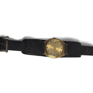 Könnte beinhalten: Eine goldfarbene Armbanduhr mit einem schwarzen Lederarmband und einer silbernen Schließe. Das Zifferblatt der Uhr ist weiß mit schwarzen römischen Ziffern als Stundenmarkierungen und schwarzen Zeigern.