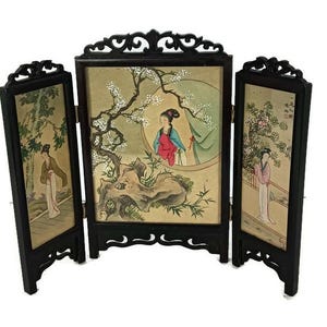 Può includere: Uno schermo in legno nero a tre pannelli con scene dipinte di donne in abiti tradizionali e motivi floreali. Lo schermo è decorato con sculture intricate.