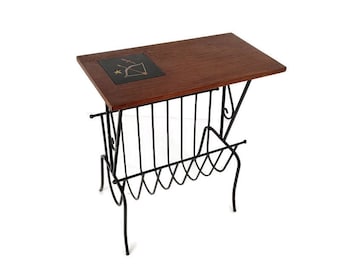 Puede incluir: Una mesa auxiliar de mediados de siglo con una tapa de madera oscura y una estructura de metal negro. La mesa tiene un revistero incorporado y una pequeña incrustación cuadrada de azulejo con un diseño de flecha y arco. La mesa mide aproximadamente 61 cm de largo, 41 cm de ancho y 51 cm de alto.