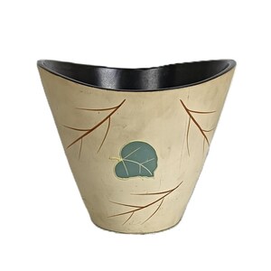 May include: A decorative, cream-colored vase with a black interior. The vase features a leaf design with a teal leaf and brown branch accents. The vase has a unique, curved shape.