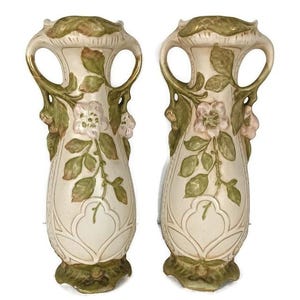 Könnte beinhalten: Paar antike Porzellanvasen mit Blumendesigns. Die Vasen sind cremefarben mit grünen und rosa floralen Akzenten. Jede Vase hat zwei Henkel und eine dekorative Basis.