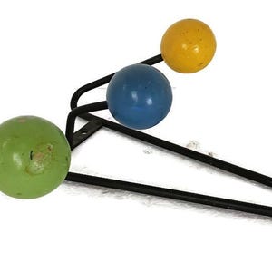 Pode incluir: Um cabide de parede moderno de meados do século. Possui uma estrutura de metal preta com três esferas de madeira coloridas em verde, azul e amarelo. As esferas são dispostas em um padrão escalonado, proporcionando uma solução de armazenamento funcional e decorativa.