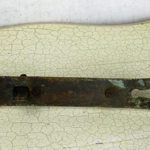 May include: An antique, weathered metal door latch with a keyhole and decorative ends. The latch is a dark, tarnished brown with hints of green and blue oxidation, and is mounted on a white, cracked surface.