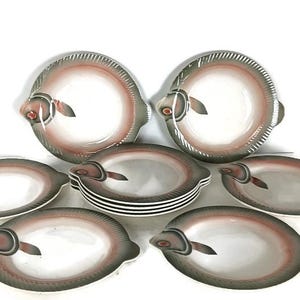 Puede incluir: Un juego de platos y fuentes de cerámica, cada uno diseñado para parecerse a un pez. Los platos son blancos con un diseño en forma de pez en tonos grises, marrones y rojos. El juego incluye platos redondos y fuentes ovaladas.