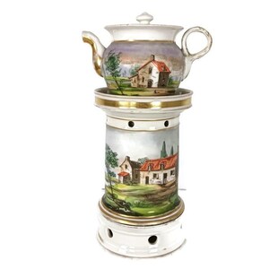Puede incluir: Una tetera de porcelana blanca con ribete dorado y una escena pintada de una casa y árboles. La tetera está sobre un soporte a juego con una escena pintada similar.