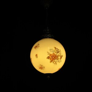 May include: A vintage, spherical pendant light with a cream-colored glass shade. The shade is adorned with floral patterns in shades of brown, orange, and green. The light fixture hangs from a decorative metal canopy and chain against a black background.