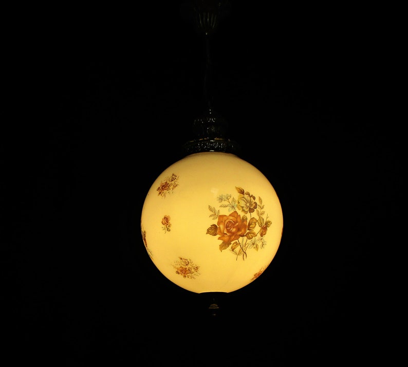 May include: A vintage, spherical pendant light with a cream-colored glass shade. The shade is adorned with floral patterns in shades of brown, orange, and green. The light fixture hangs from a decorative metal canopy and chain against a black background.
