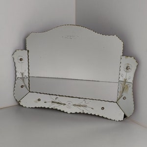 Op de afbeelding: Een vintage spiegel met een afgeschuinde rand en een decoratief bloemmotief. De spiegel heeft een gegolfde rand en is ingelijst in een zilverkleurig metaal.