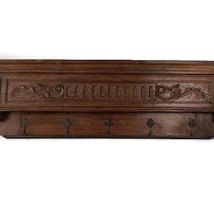 May include: A dark brown wooden wall shelf with decorative carvings and four metal hooks. The shelf has a detailed design with floral and geometric patterns, suitable for entryway or hallway decor.