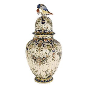 Può includere: Un vaso decorativo in ceramica color crema con coperchio e figura di uccello. Presenta intricati motivi floreali e fogliari in blu, verde e arancione. Il vaso ha una texture a coste che ne esalta il design.