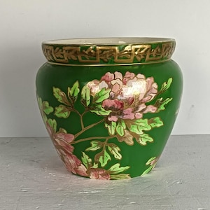 Peut inclure: Un pot en céramique vert avec un motif floral. Le pot a un bord doré et une bande décorative autour du haut. Le motif floral présente des fleurs roses et blanches avec des feuilles vertes et des accents dorés. Le pot est probablement utilisé à des fins décoratives.