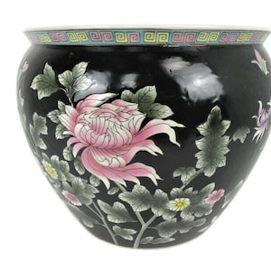 Peut inclure: Pot de fleurs en céramique noire de forme arrondie. Il est décoré d'une grande fleur rose et blanche, de feuilles vertes et de petites fleurs. Une bordure géométrique colorée entoure le bord. Des papillons sont également peints sur le pot.