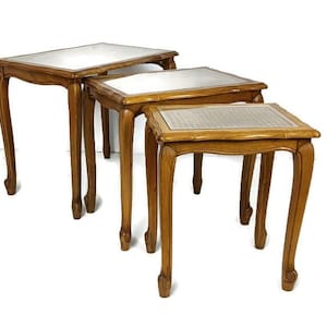 Puede incluir: Juego de tres mesas nido de color marrón claro. Cada mesa tiene una tapa rectangular con una inserción de vidrio transparente y patas curvas. La mesa más pequeña mide unos 46 cm de ancho.