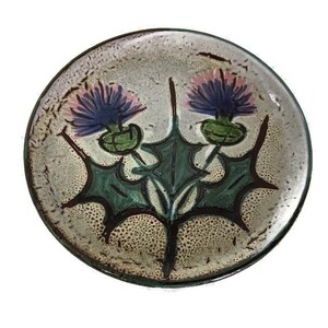 May include: Round ceramic plate with a speckled, off-white background. The plate features a central design of two purple thistle flowers with green leaves and stems. The rim is a darker green.