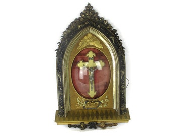 May include: An antique religious wall hanging featuring a crucifix. The ornate frame is gold and dark bronze, with a red velvet background. The cross has a silver figure of Jesus. The top of the frame has a gold sunburst design.