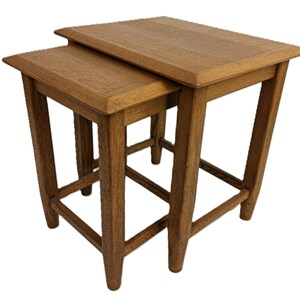 Peut inclure: Un ensemble de deux tables gigognes en bois clair. Les tables présentent un grain de bois naturel et des pieds robustes et inclinés. Les plateaux ont un bord surélevé. La petite table est conçue pour s'emboîter sous la plus grande. Idéal pour un salon.