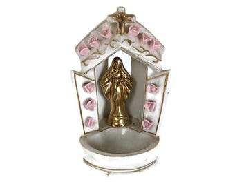 May include: A white ceramic holy water font with a gold-colored statue of the Virgin Mary. The font is decorated with pink roses and gold accents. A cross is at the top of the structure.