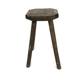 Puede incluir: Un taburete de madera marrón oscuro con tres patas y un asiento octogonal. El taburete tiene un aspecto rústico y envejecido, con vetas de madera visibles y ligeras imperfecciones. Las patas están anguladas para mayor estabilidad.