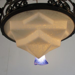 May include: An ornate, Art Deco-style chandelier with a geometric, frosted glass shade. The fixture has a black metal frame with decorative scrollwork. A small, blue glass element hangs below.
