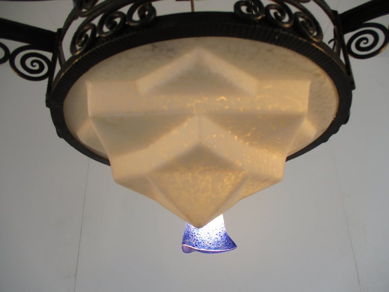 May include: An ornate, Art Deco-style chandelier with a geometric, frosted glass shade. The fixture has a black metal frame with decorative scrollwork. A small, blue glass element hangs below.